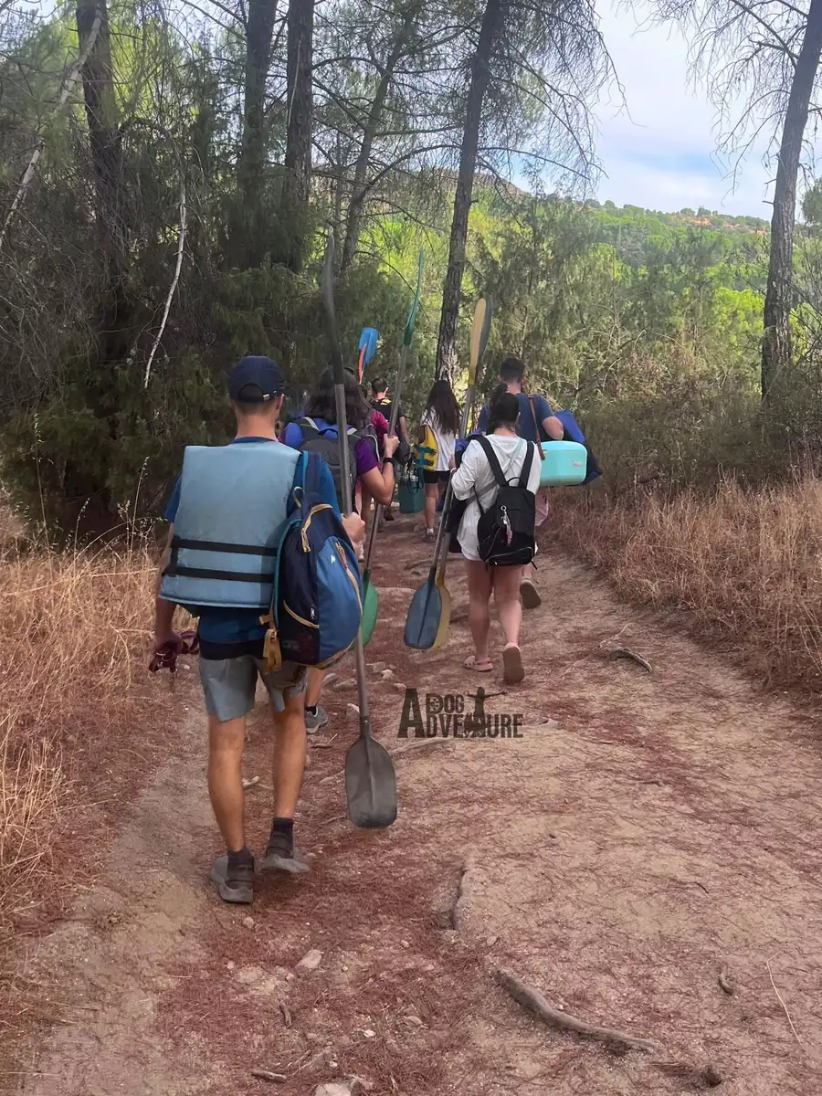Grupo caminando de espaldas por un sendero hacia el embalse con remos y chalecos